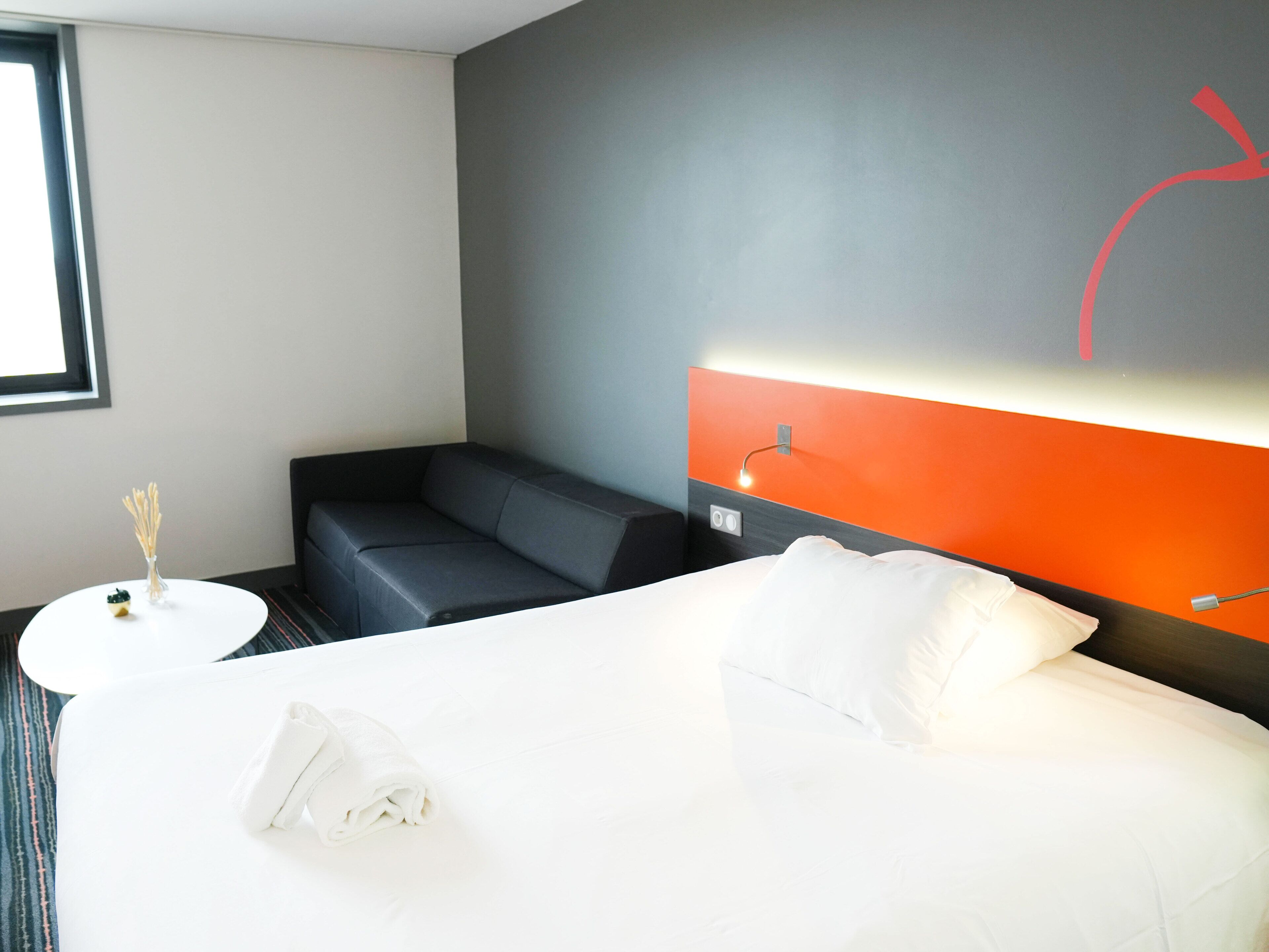 Quarto de hotel com uma cama grande, um sofá pequeno, uma mesa redonda branca e um candeeiro fixado na parede.