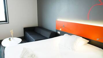 Quarto de hotel com uma cama grande, um sofá pequeno, uma mesa redonda branca e um candeeiro fixado na parede.