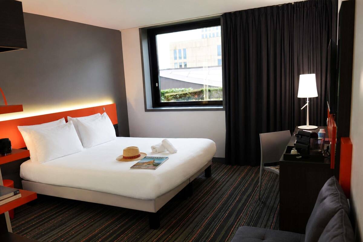 Hotelkamer met een bed, bureau, stoel, raam met gordijnen en een lamp.