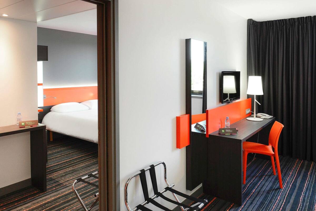 Een hotelkamer met een bed, een bureau met stoel, een spiegel, een televisie en een raam met gordijnen.