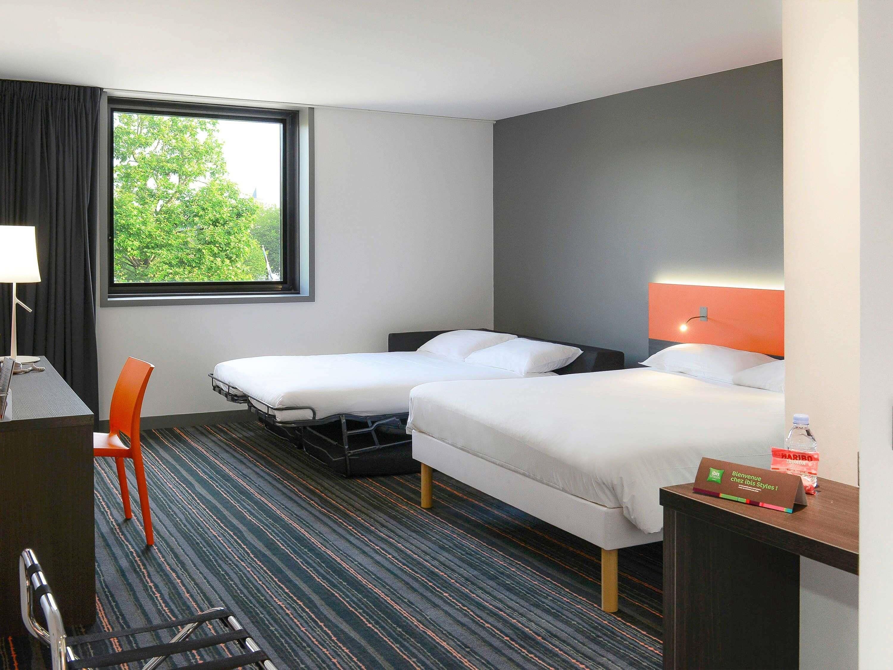 A hotel room with two beds, a desk, a chair, a window with a view of trees, and a sign on the desk.