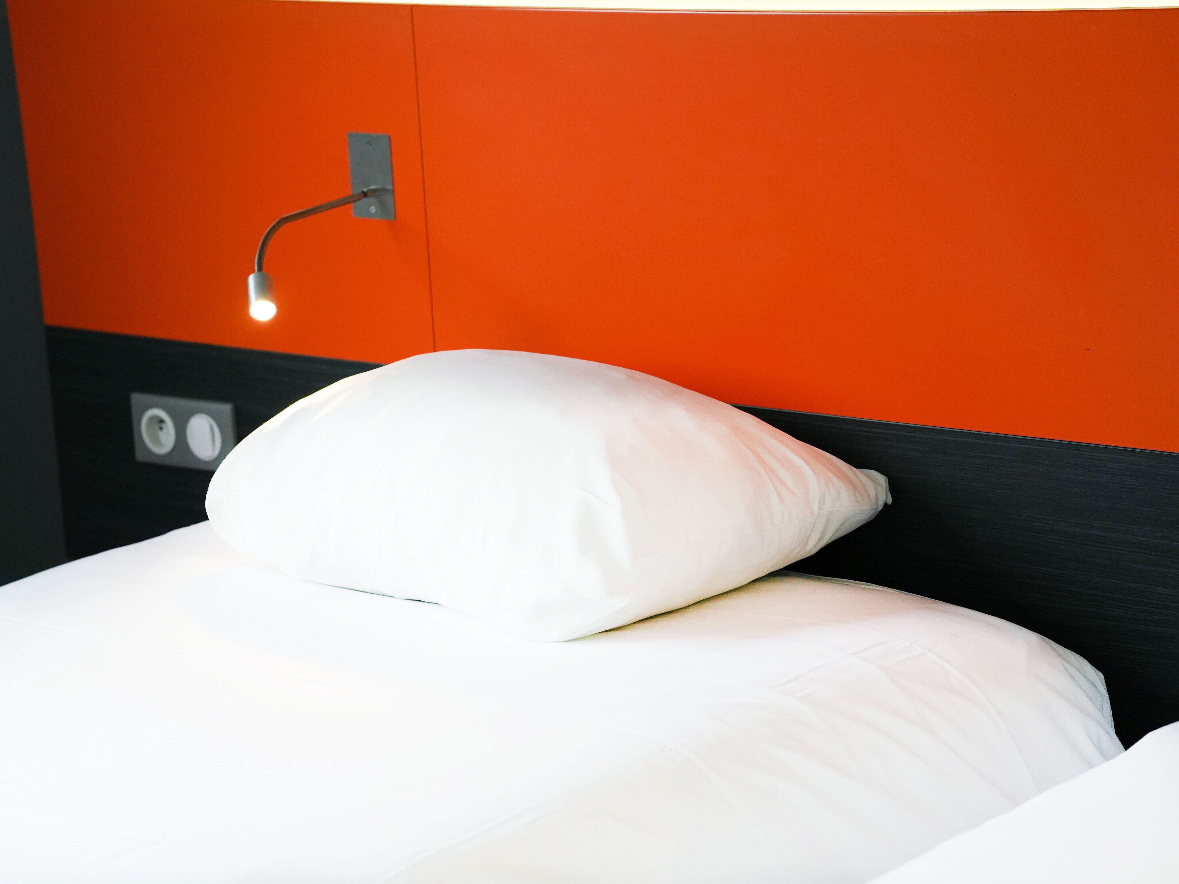 A bed with white bedding and a white pillow, a wall socket, and a light fixture.