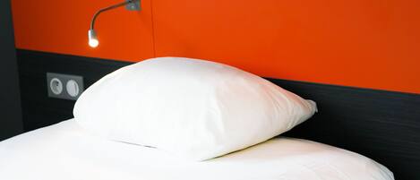 A bed with white bedding and a white pillow, a wall socket, and a light fixture.
