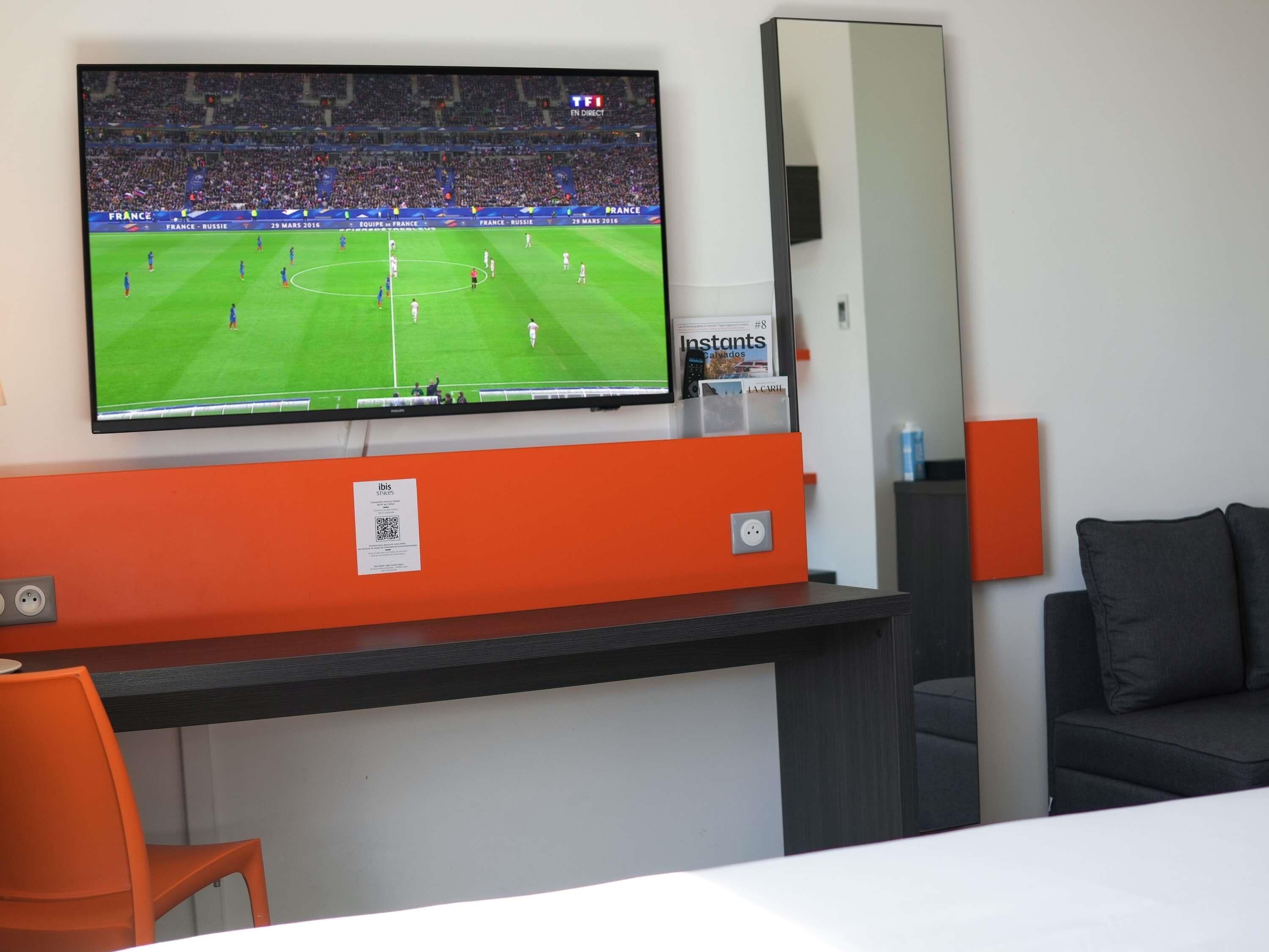 A hotel room with a TV mounted on the wall displaying a soccer match, a desk with a mirror, and a couch.