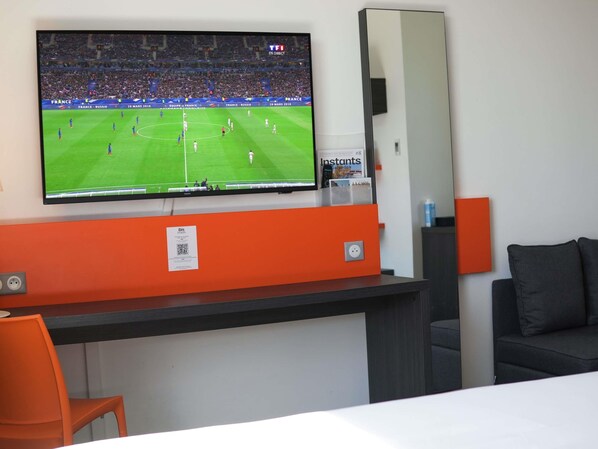 A hotel room with a TV mounted on the wall displaying a soccer match, a desk with a mirror, and a couch. - Ibis Styles Caen Centre Gare (Caen)