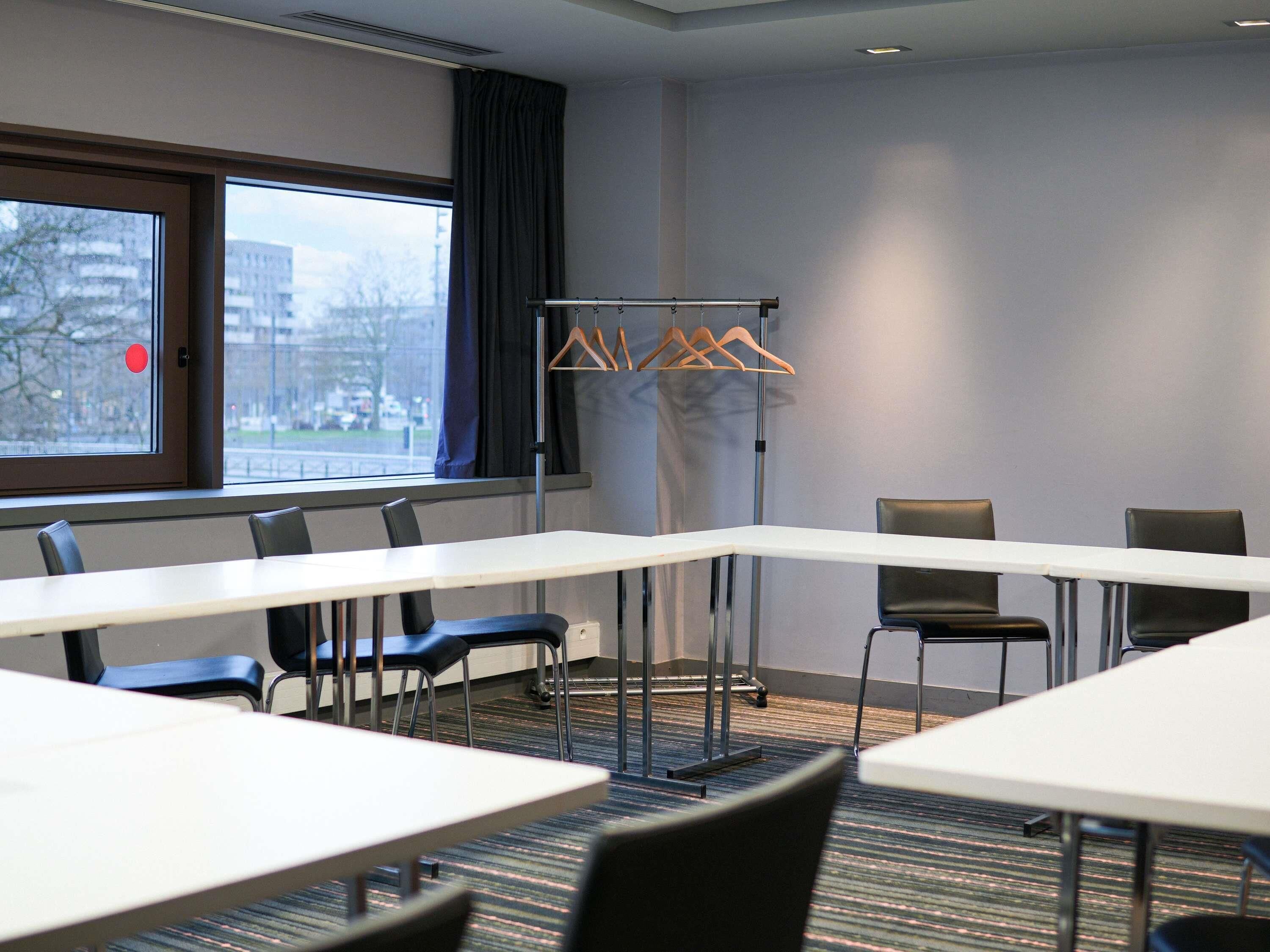 Une salle de conférence avec une longue table, des chaises, un porte-manteau et une fenêtre donnant sur des immeubles.