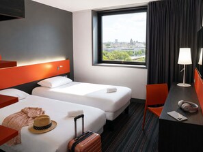 A hotel room with two beds, a view of the city, a desk with a lamp, and a suitcase. - Ibis Styles Caen Centre Gare (Caen)
