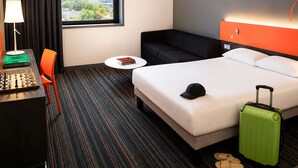 Habitación de hotel con cama, escritorio con lámpara, una silla, una mesita, sofá y vistas a la ciudad.