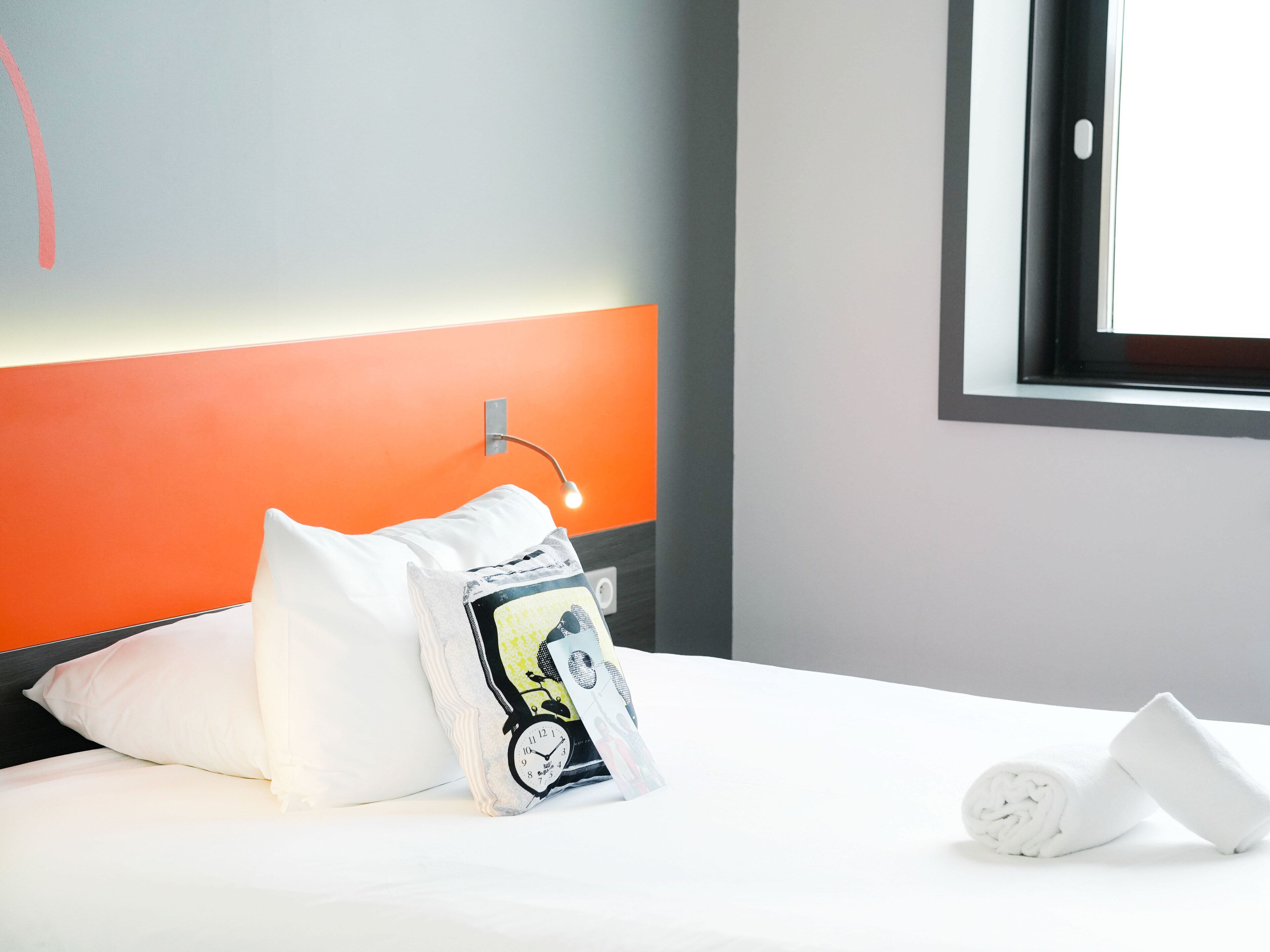 A neatly made bed with white linens, a pillow, and a rolled towel.