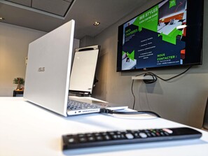 A laptop on a desk with a TV displaying a presentation, a remote control, and a plant in the background. - Ibis Styles Caen Centre Gare (Caen)
