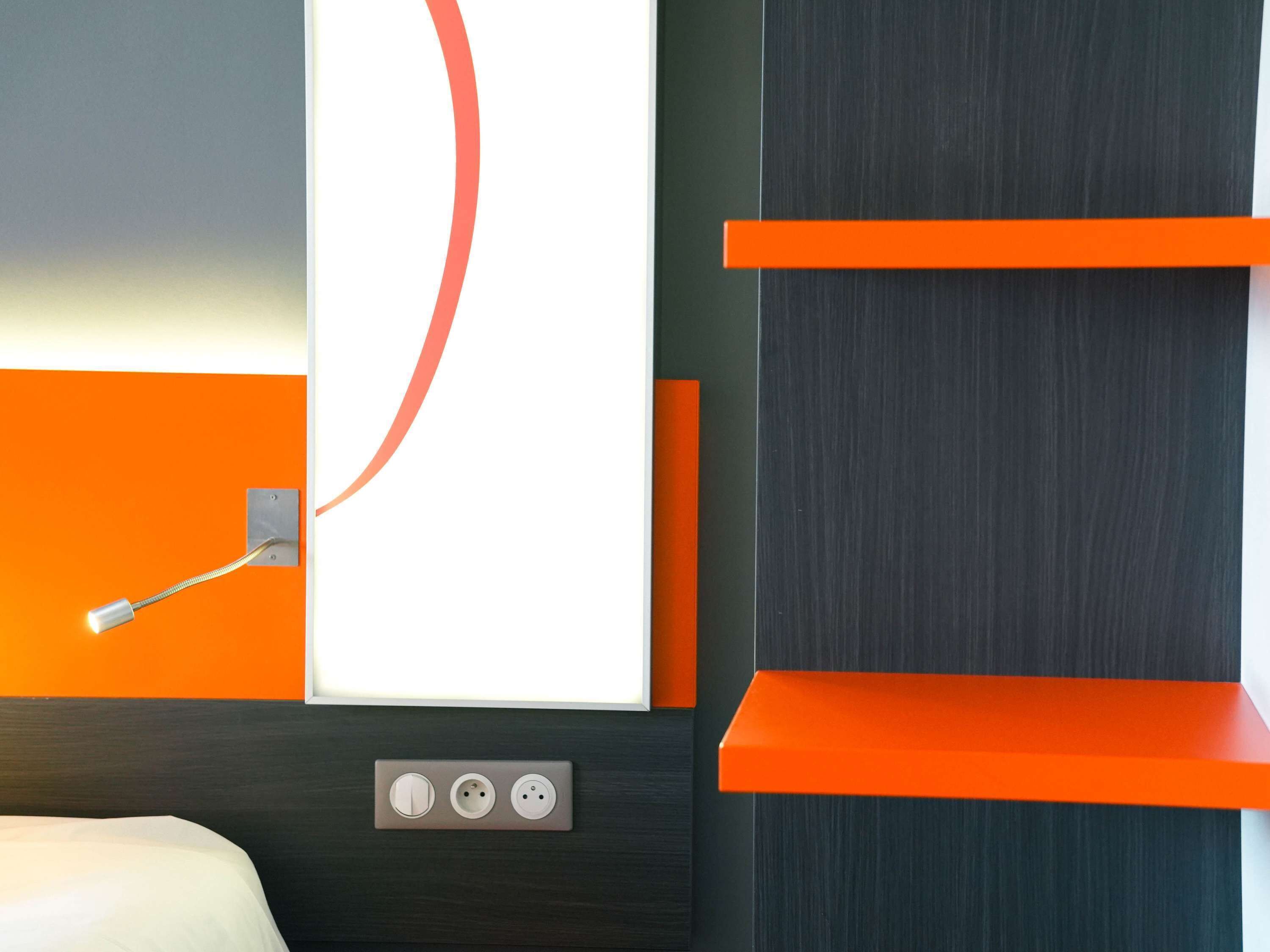 A modern hotel room with a bed, a wall-mounted light fixture, and a power outlet with three sockets.