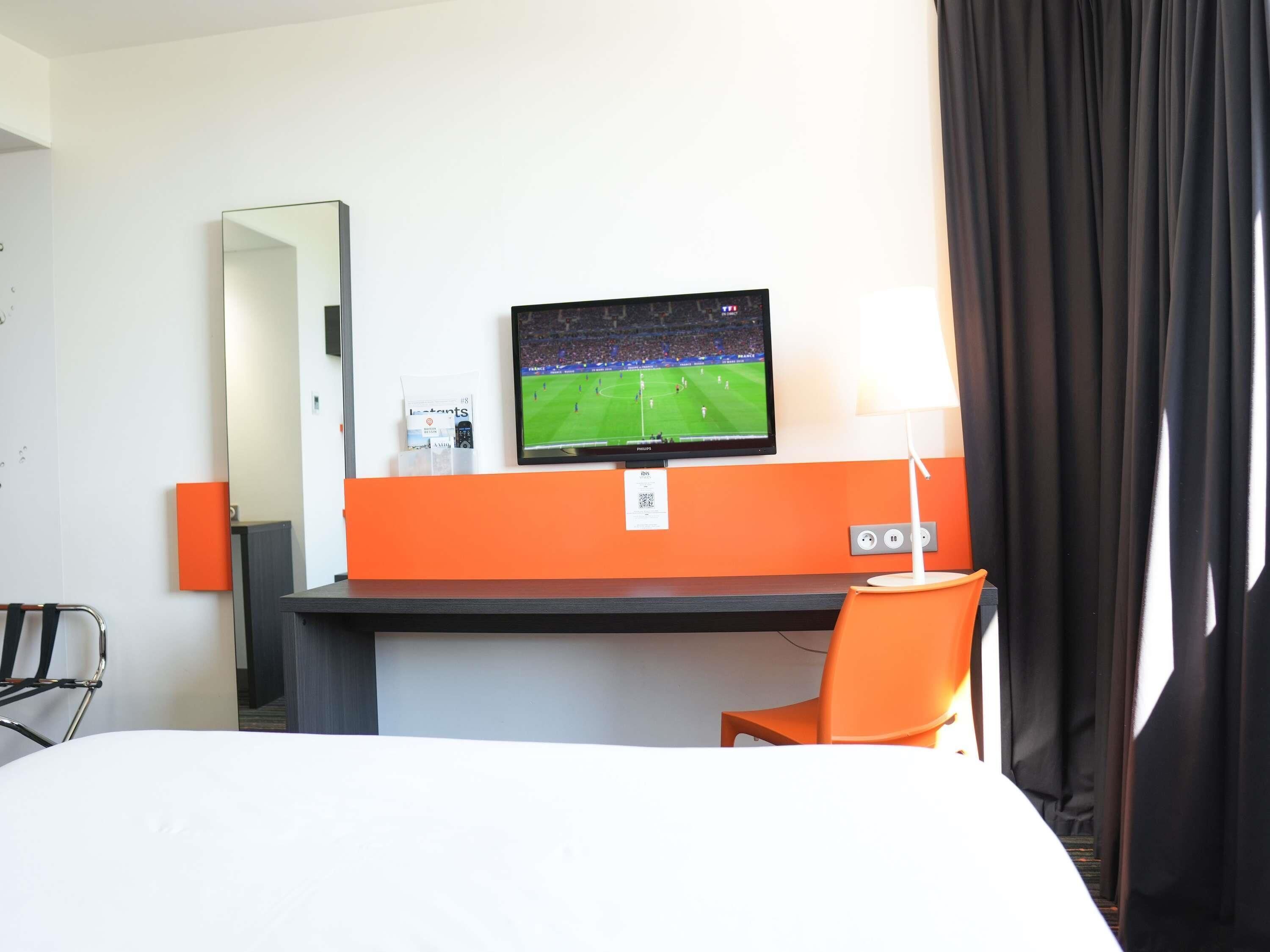 Habitación de hotel con cama, escritorio con televisor, silla, espejo y ventana con cortinas.