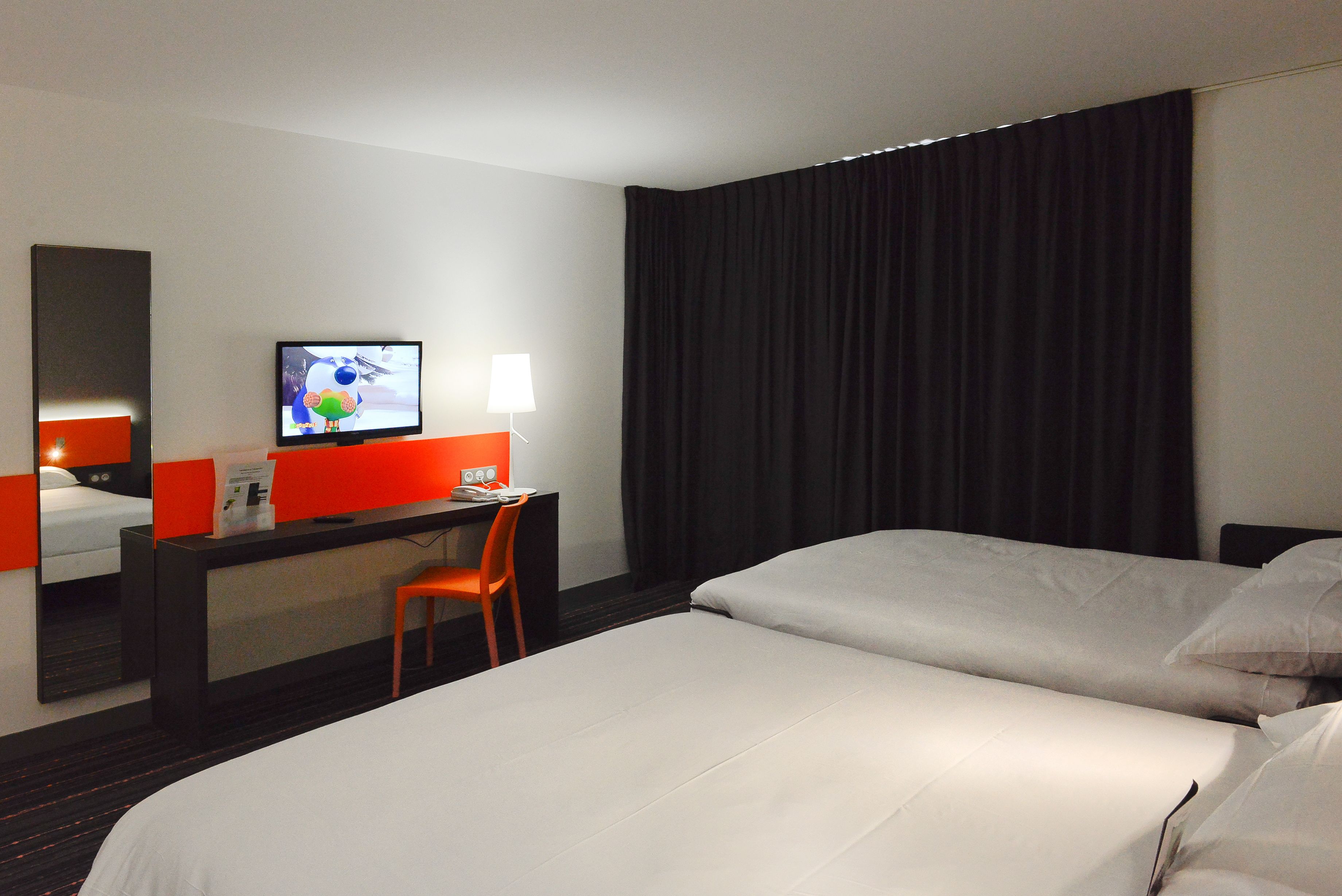 A hotel room with a bed, a desk with a chair, a television, and a window with curtains.