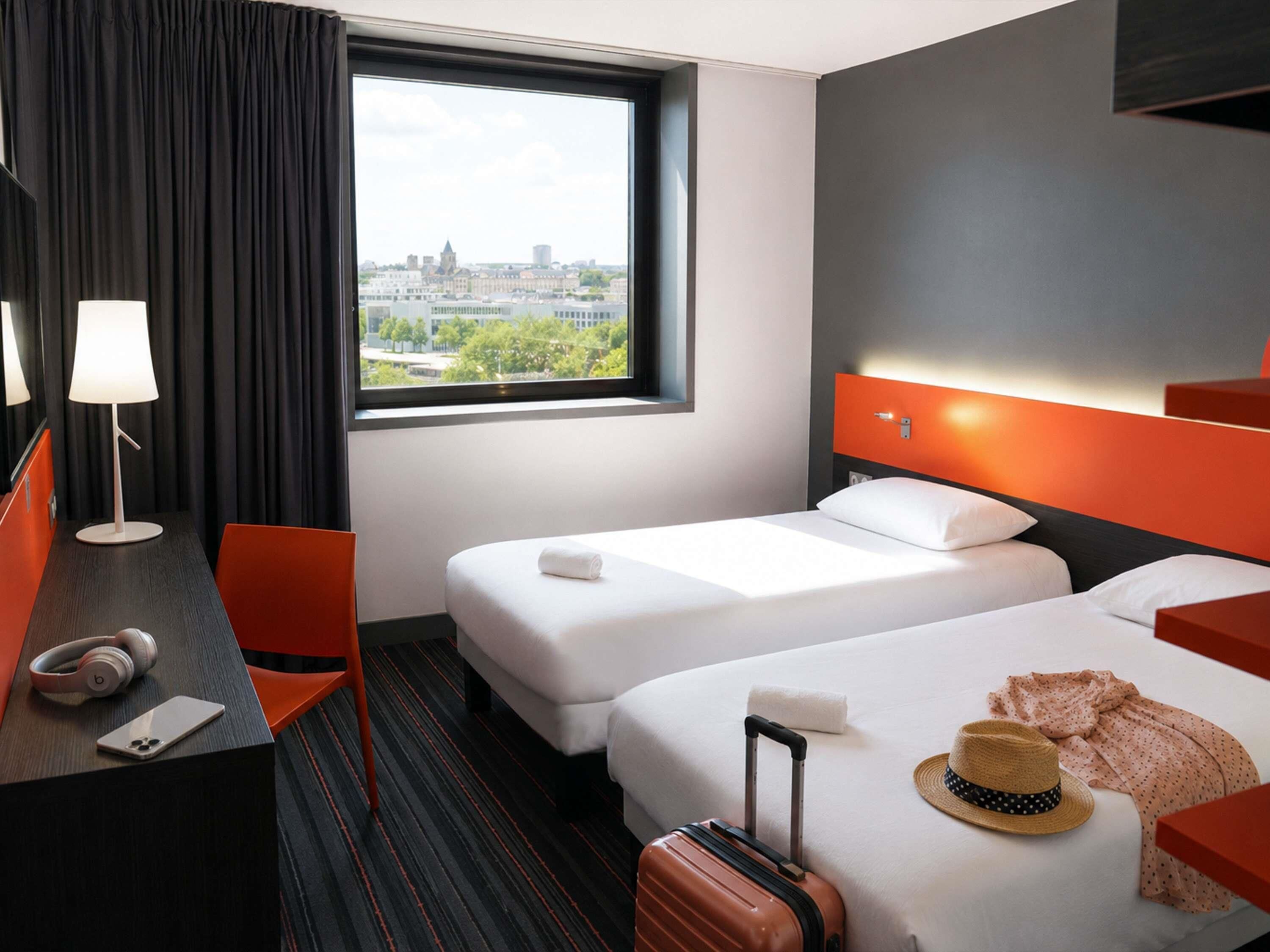 Habitación de hotel con dos camas, un escritorio con lámpara, una silla, una maleta y vistas a la ciudad.