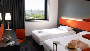 Habitación de hotel con dos camas, un escritorio con lámpara, una silla, una maleta y vistas a la ciudad.
