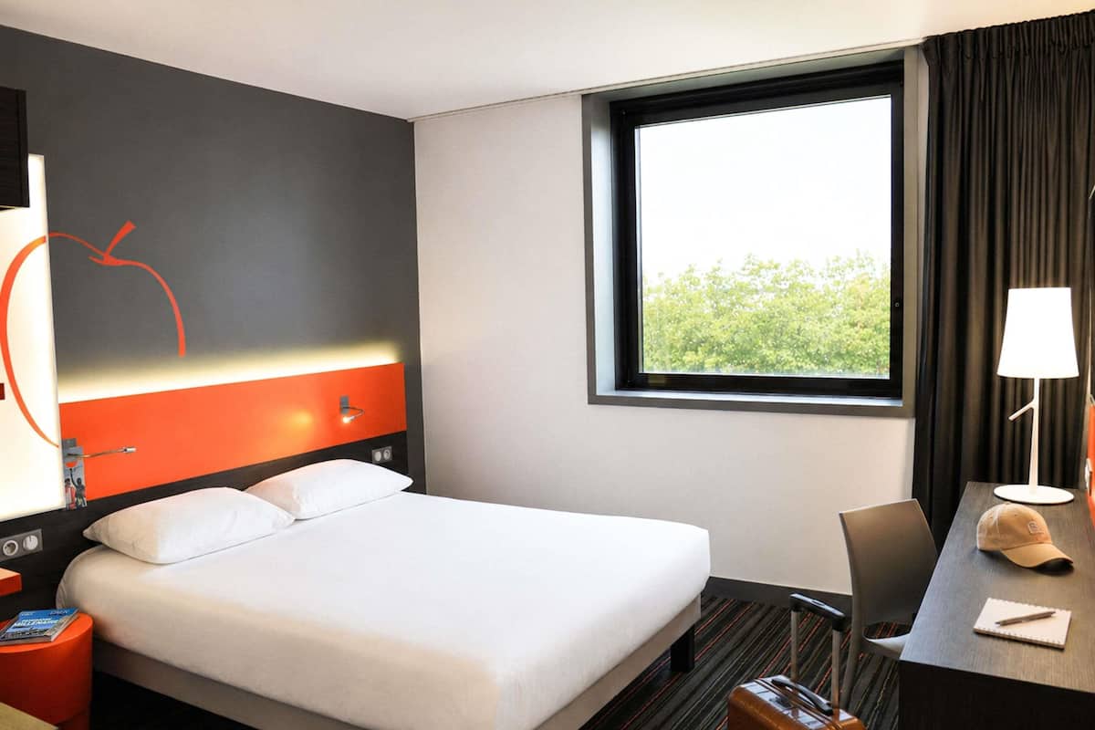 Hotelkamer met een bed, een bureau, een stoel, een lamp, een koffer en een raam met uitzicht op groen.