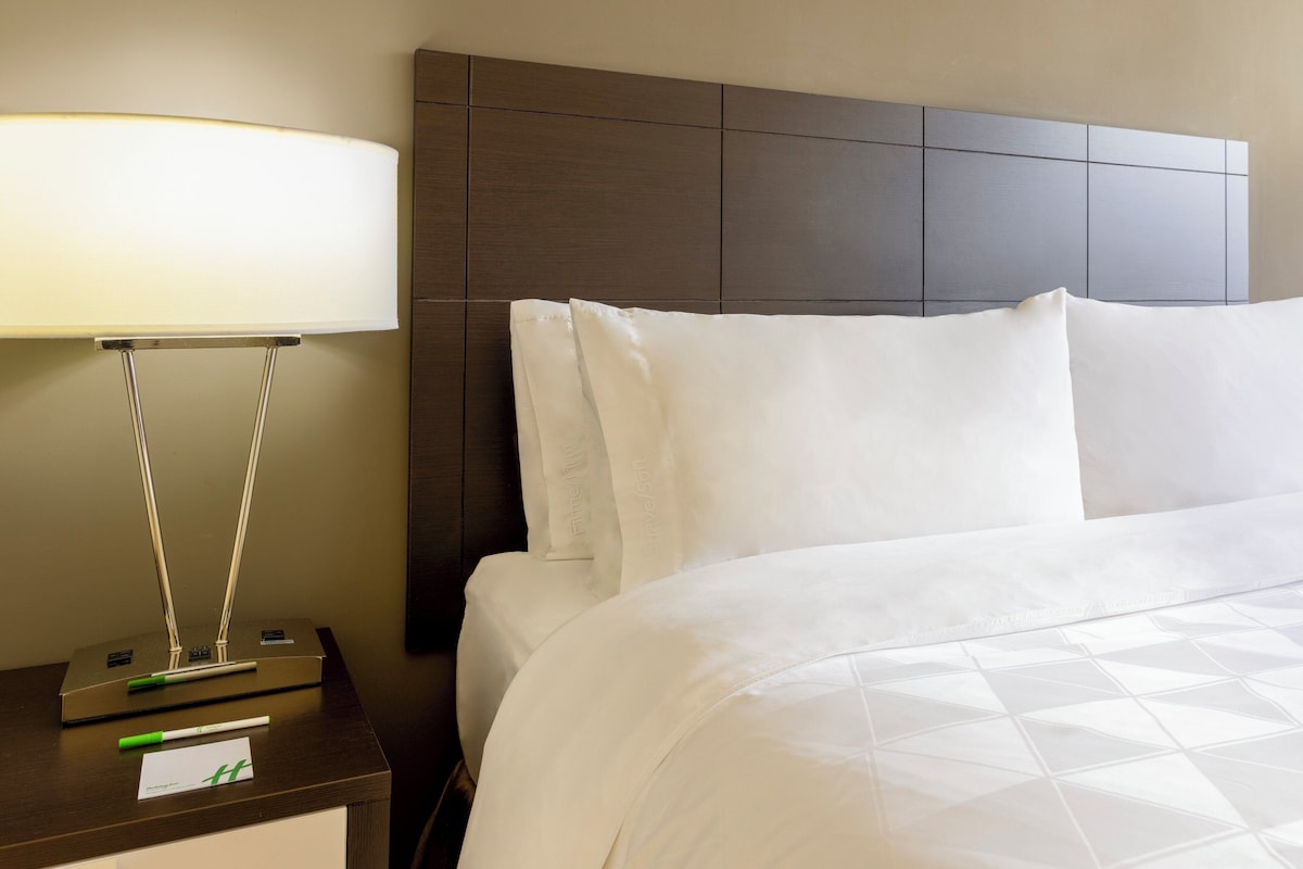 Uma cama bem arrumada com lençóis brancos, uma luminária moderna em uma mesa de cabeceira e uma cabeceira com padrão geométrico.