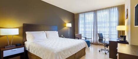 Habitación de hotel con una cama grande, un escritorio, una silla y un ventanal con cortinas.
