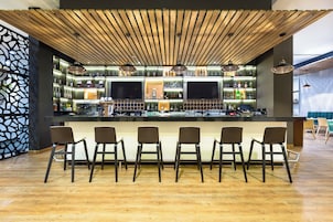 Uma área de bar com teto de madeira, várias prateleiras com garrafas e banquetas.