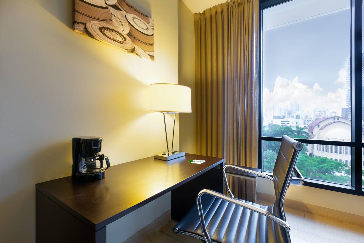 A hotel room with a desk, a chair, a coffee maker, a lamp, a window with curtains, and a cityscape view.