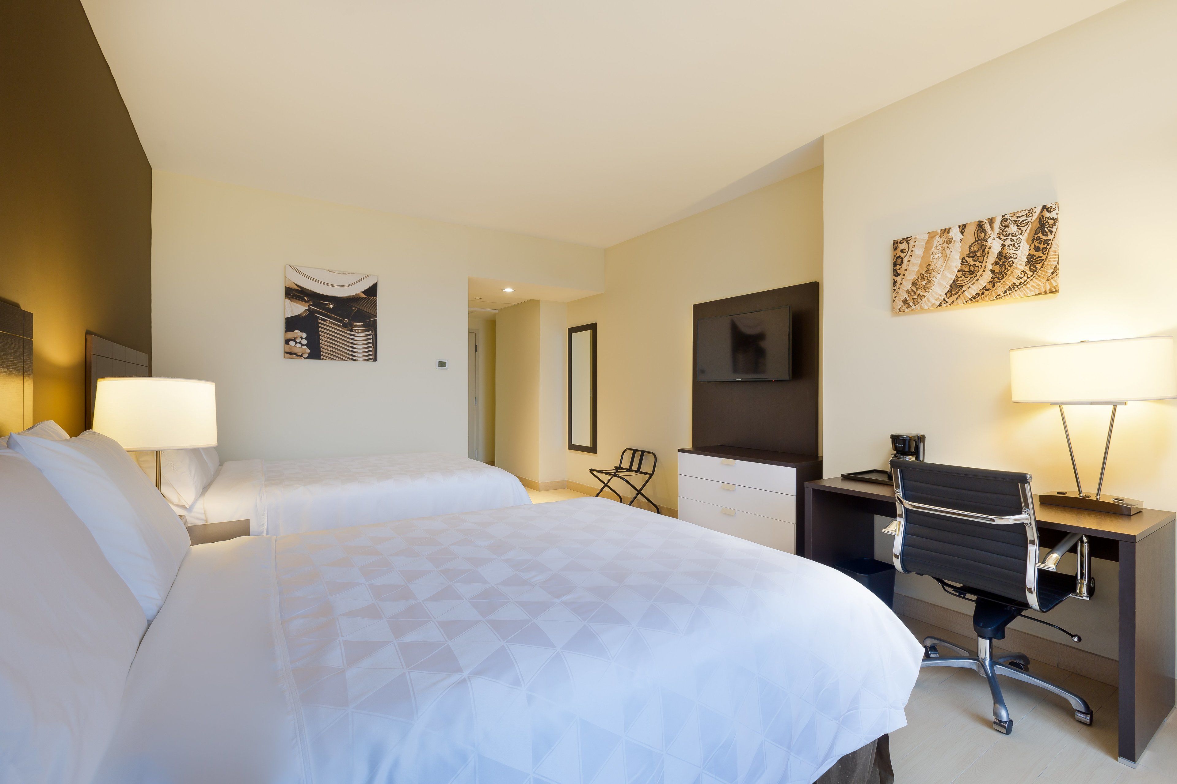 Habitación de hotel con dos camas, un escritorio, una silla, un televisor y obras de arte en las paredes.