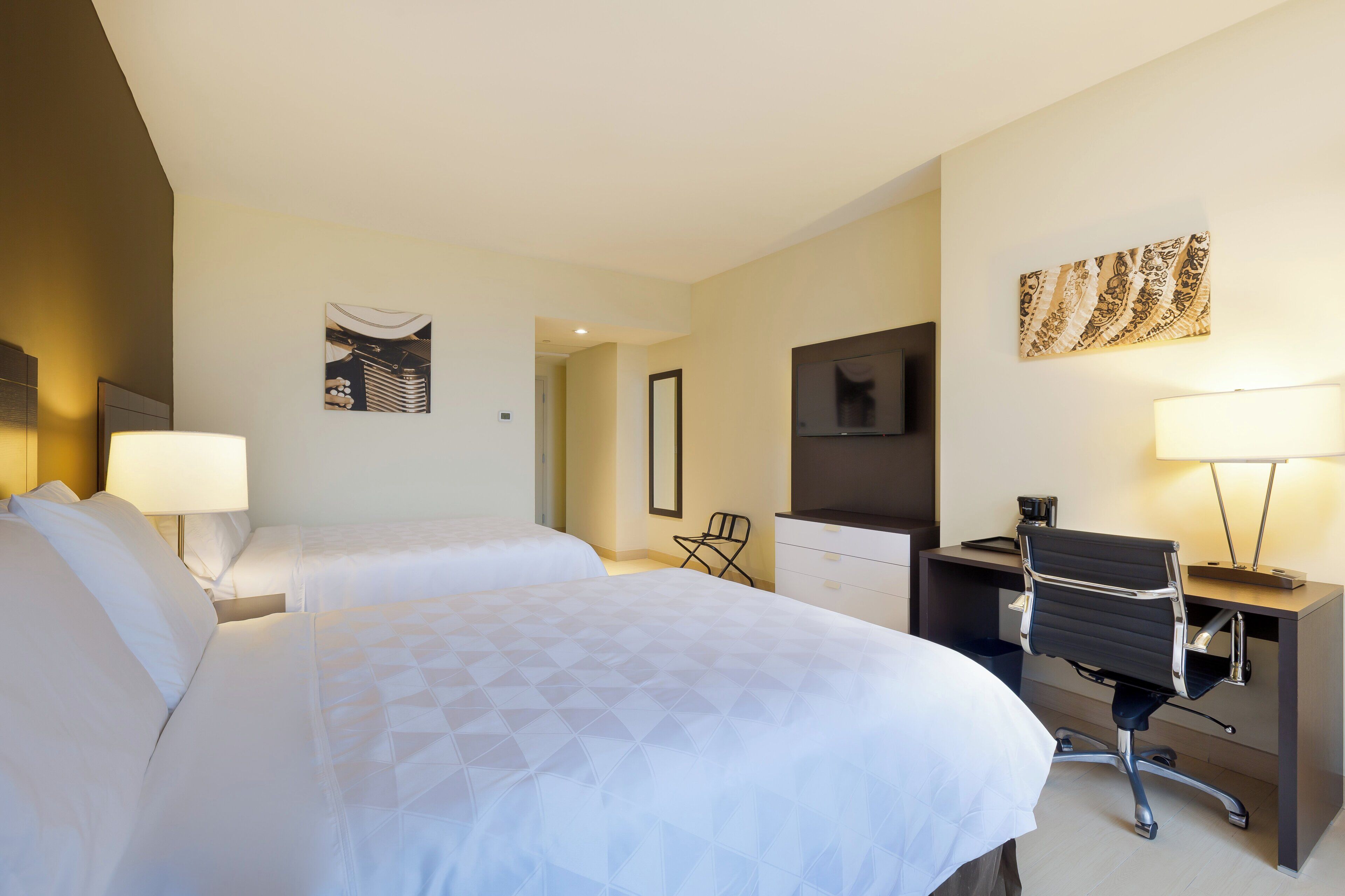 A hotel room with two beds, a desk with a chair, a television, and artwork on the walls.