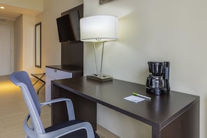 Quarto de hotel com escrivaninha, cadeira, televisão na parede, luminária e cafeteira.