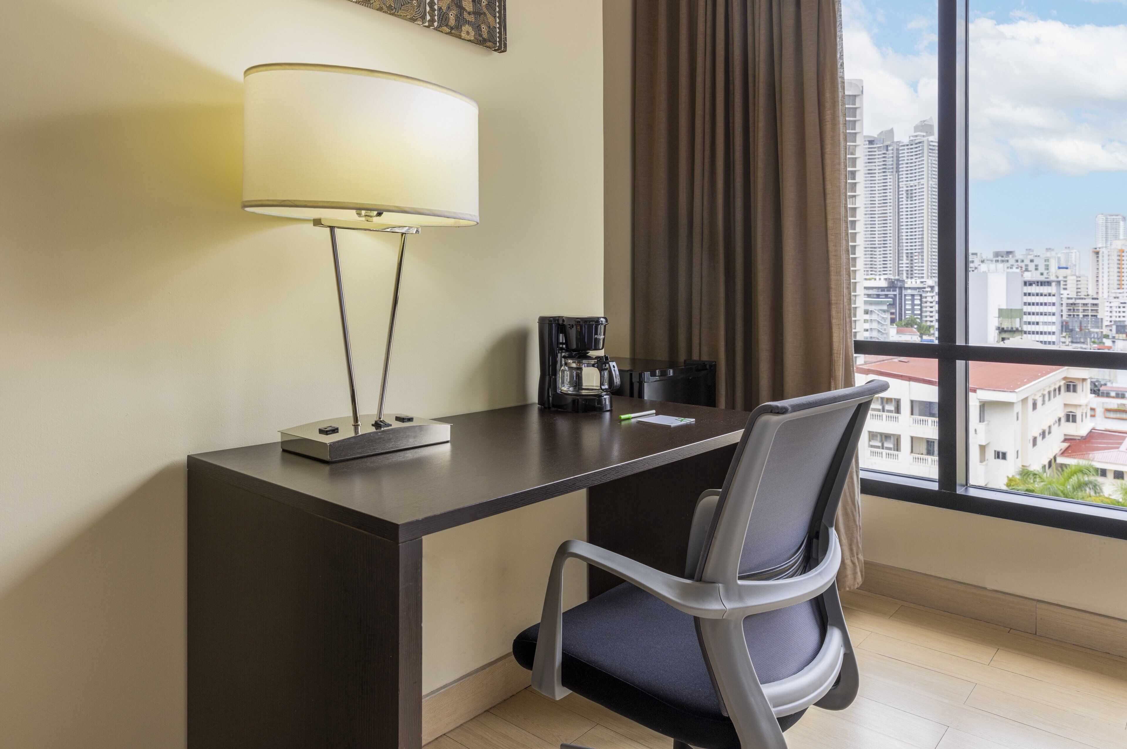 A hotel room with a desk, a lamp, a chair, a coffee maker, and a view of the cityscape through large windows.