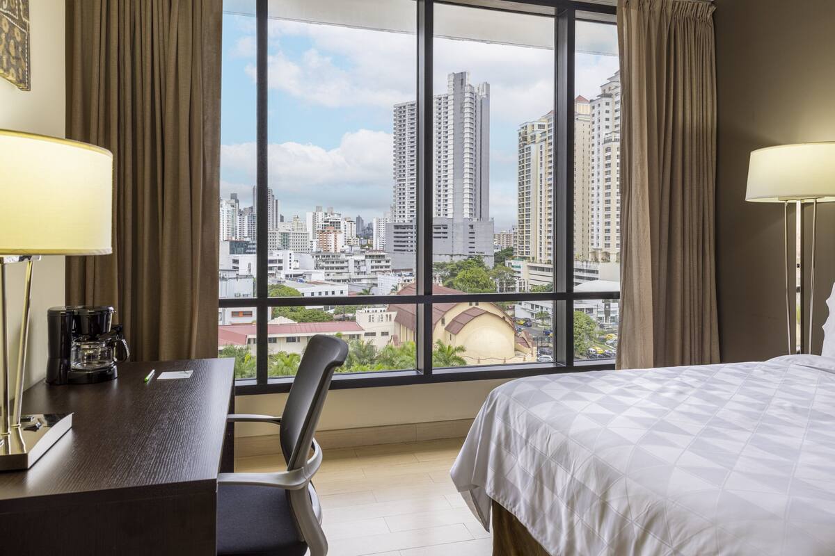 A hotel room with a large window, a bed, a desk with a coffee maker, and a chair.