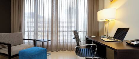 A hotel room with a bed, a desk with a laptop, a chair, a small table, and a lamp.