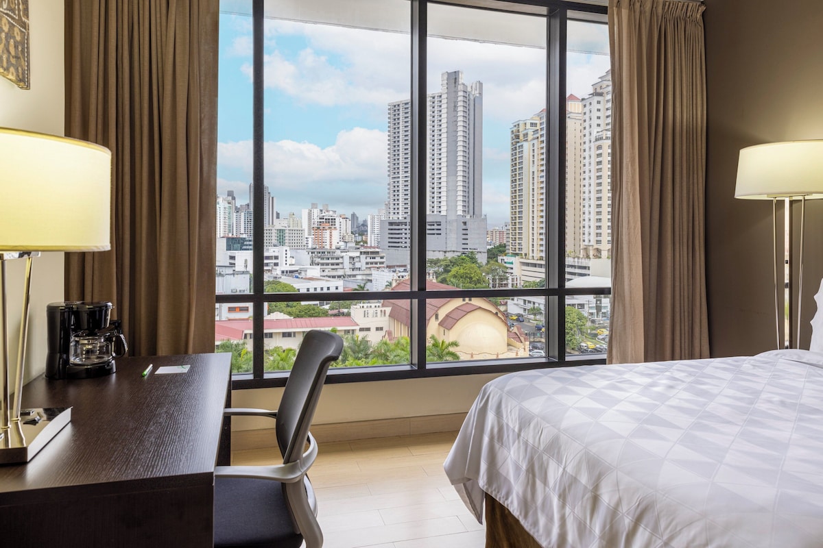 Quarto de hotel com uma janela grande, uma cama, uma escrivaninha com cafeteira e uma cadeira.