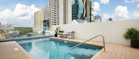 A rooftop swimming pool with a view of high-rise buildings, a wooden deck, and a seating area with chairs and a table.