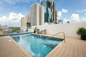 A rooftop swimming pool with a view of high-rise buildings, a wooden deck, and a seating area with chairs and a table.
