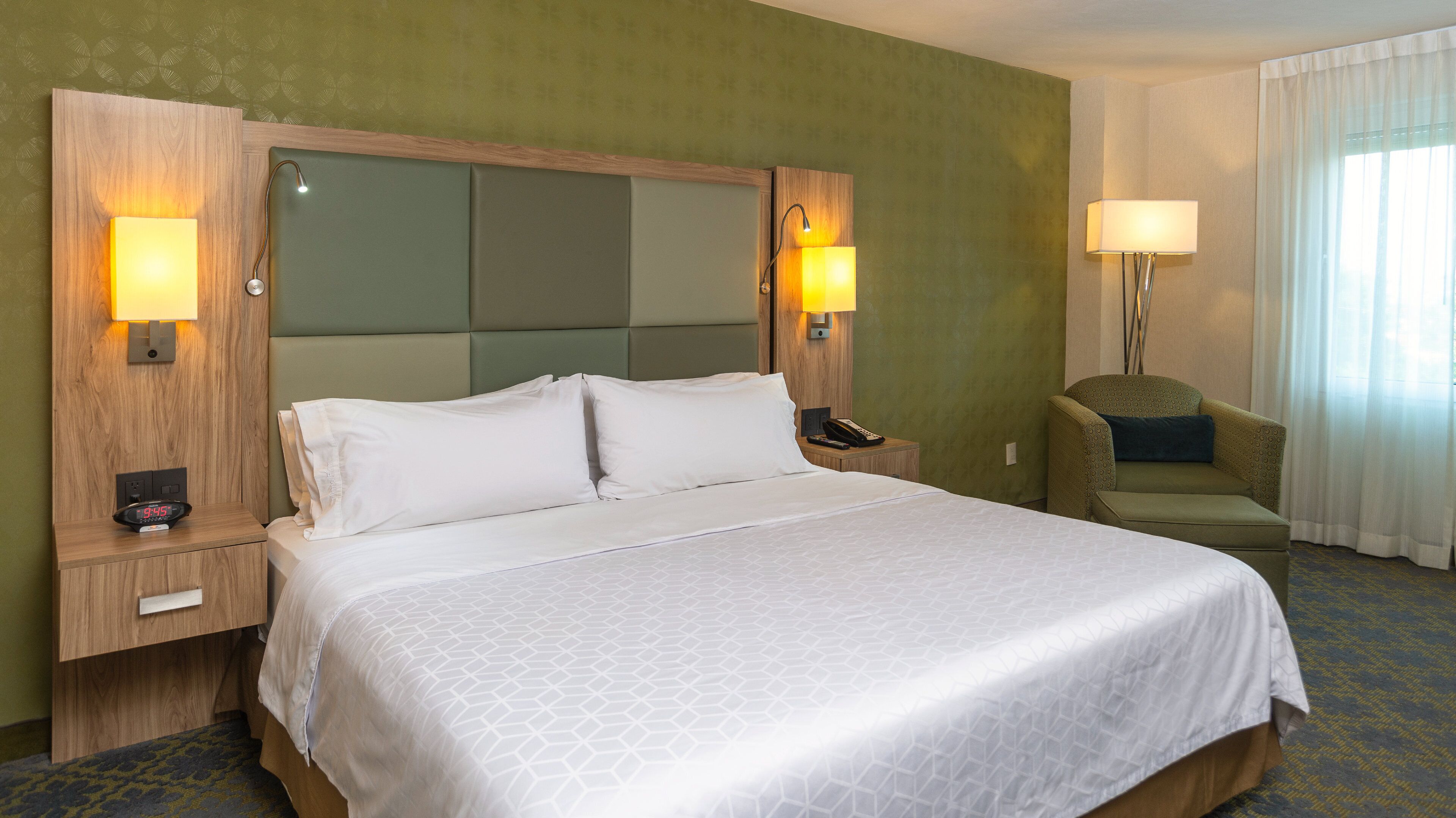 Una habitación de hotel con una cama grande, dos mesitas de noche, una silla y una lámpara.