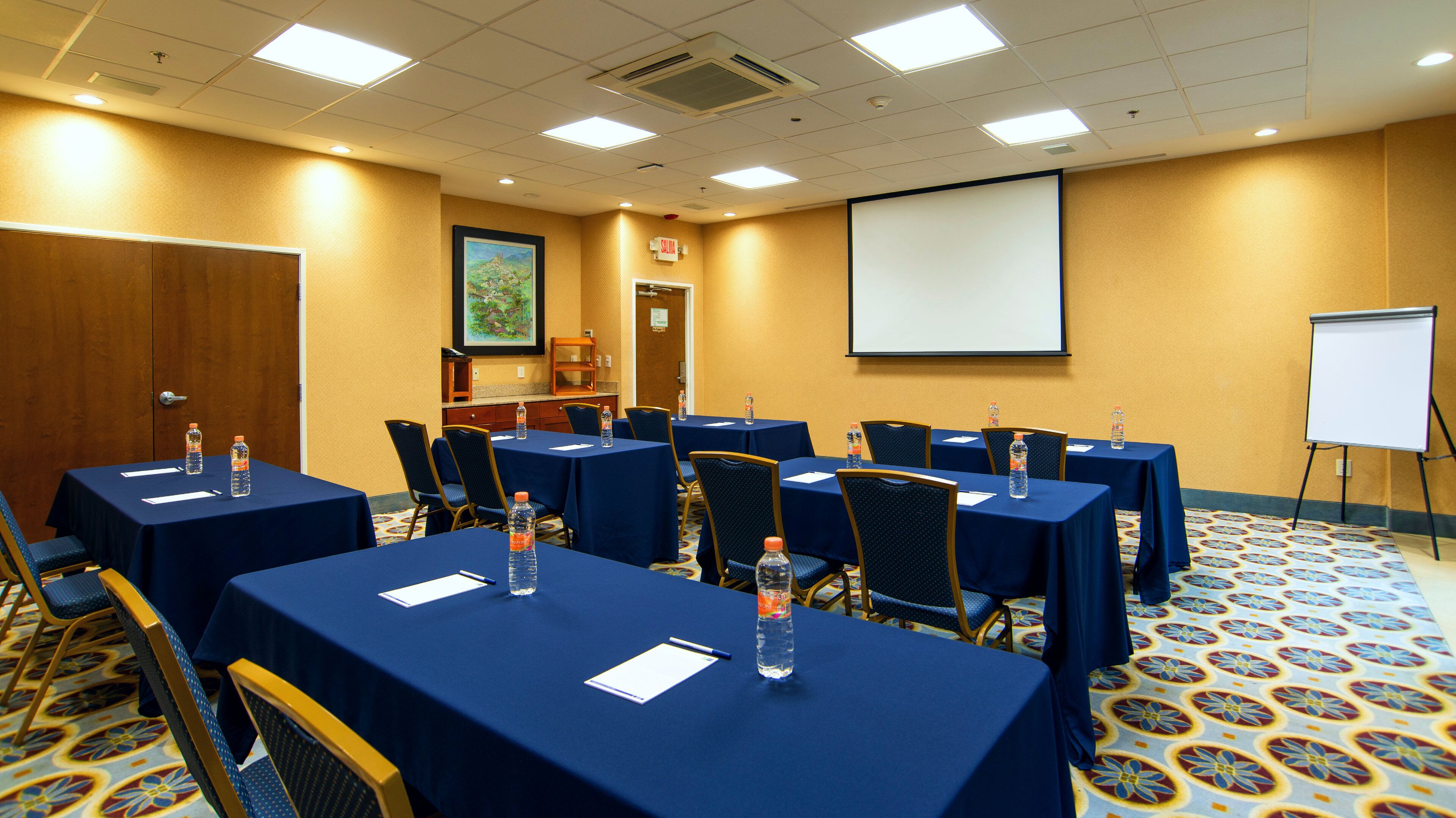 Un salón de conferencias con mesas, sillas, una pantalla de proyector y una botella de agua en cada mesa.