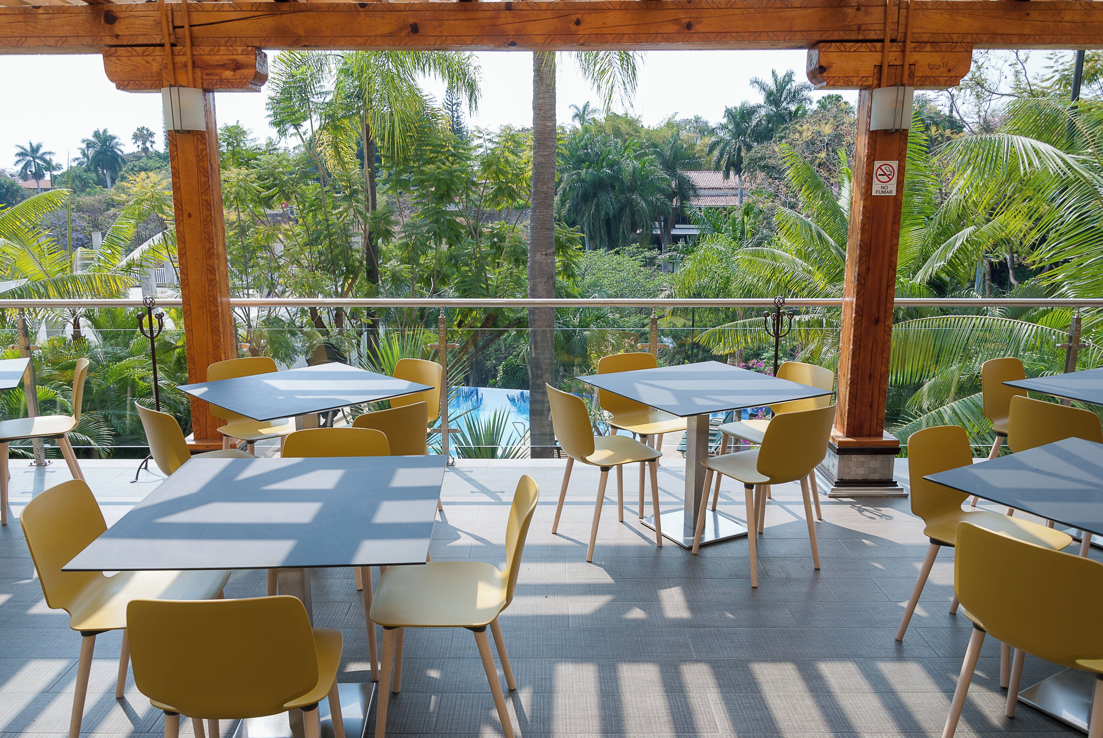 Un patio al aire libre con mesas y sillas, rodeado de abundante vegetación y vistas a una piscina.