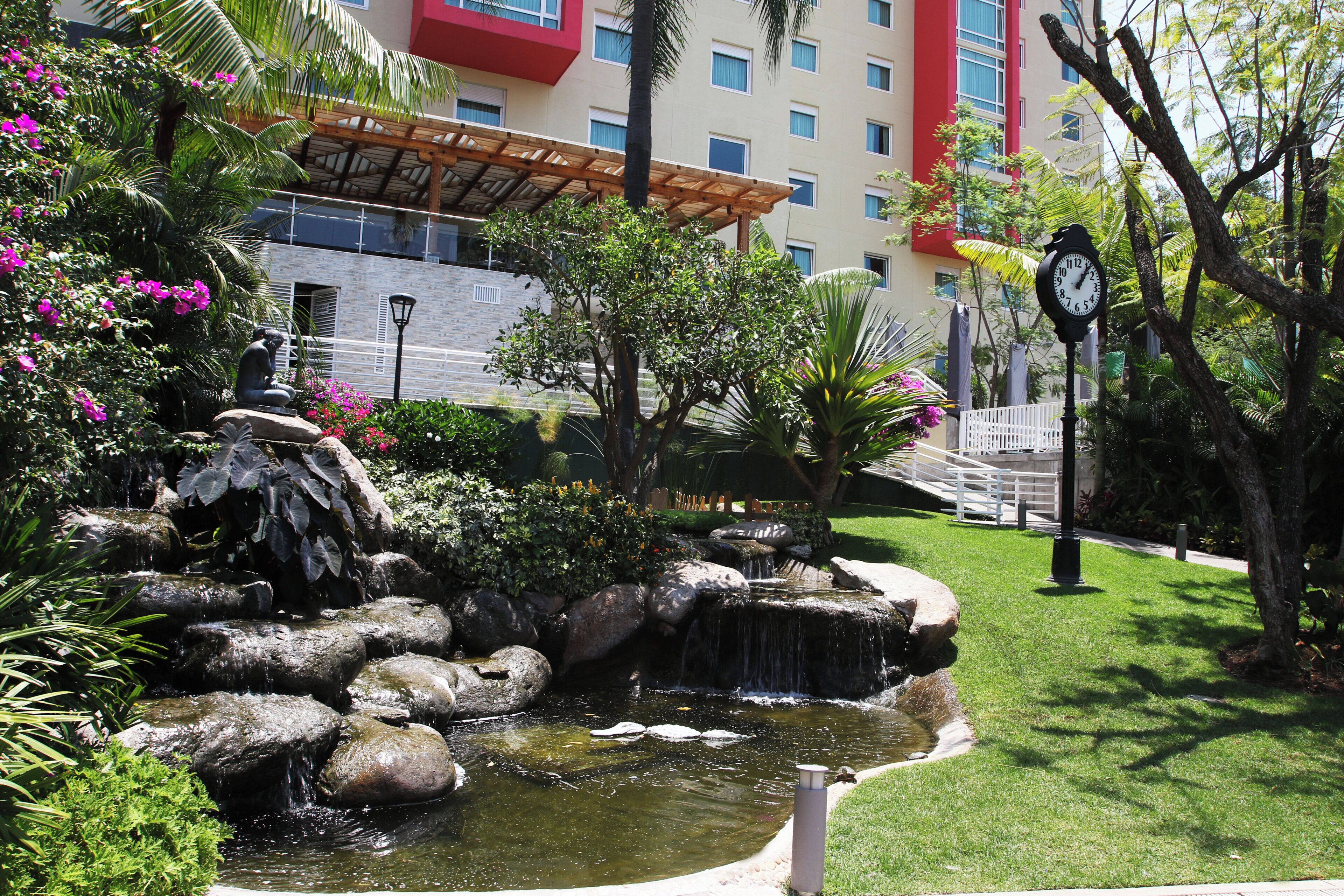 Un jardín ajardinado con fuente de agua, palmeras y un edificio de fachada roja al fondo.
