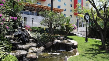 Un jardín ajardinado con fuente de agua, palmeras y un edificio de fachada roja al fondo.