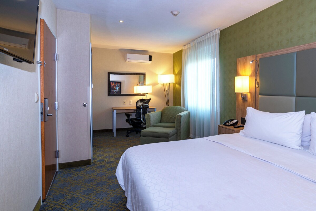 A hotel room with a bed, desk, chair, and a small table with a lamp.