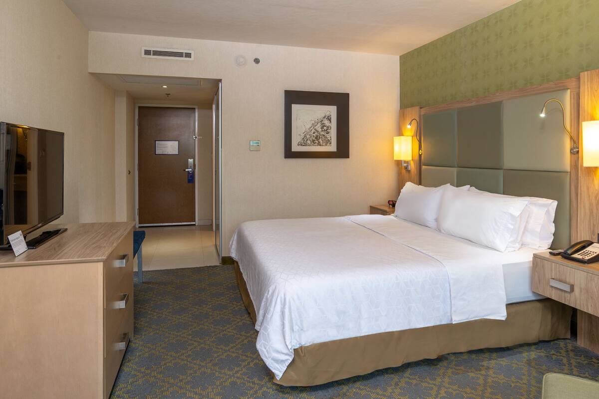 A hotel room with a bed, bedside table, lamp, and a television on a desk.