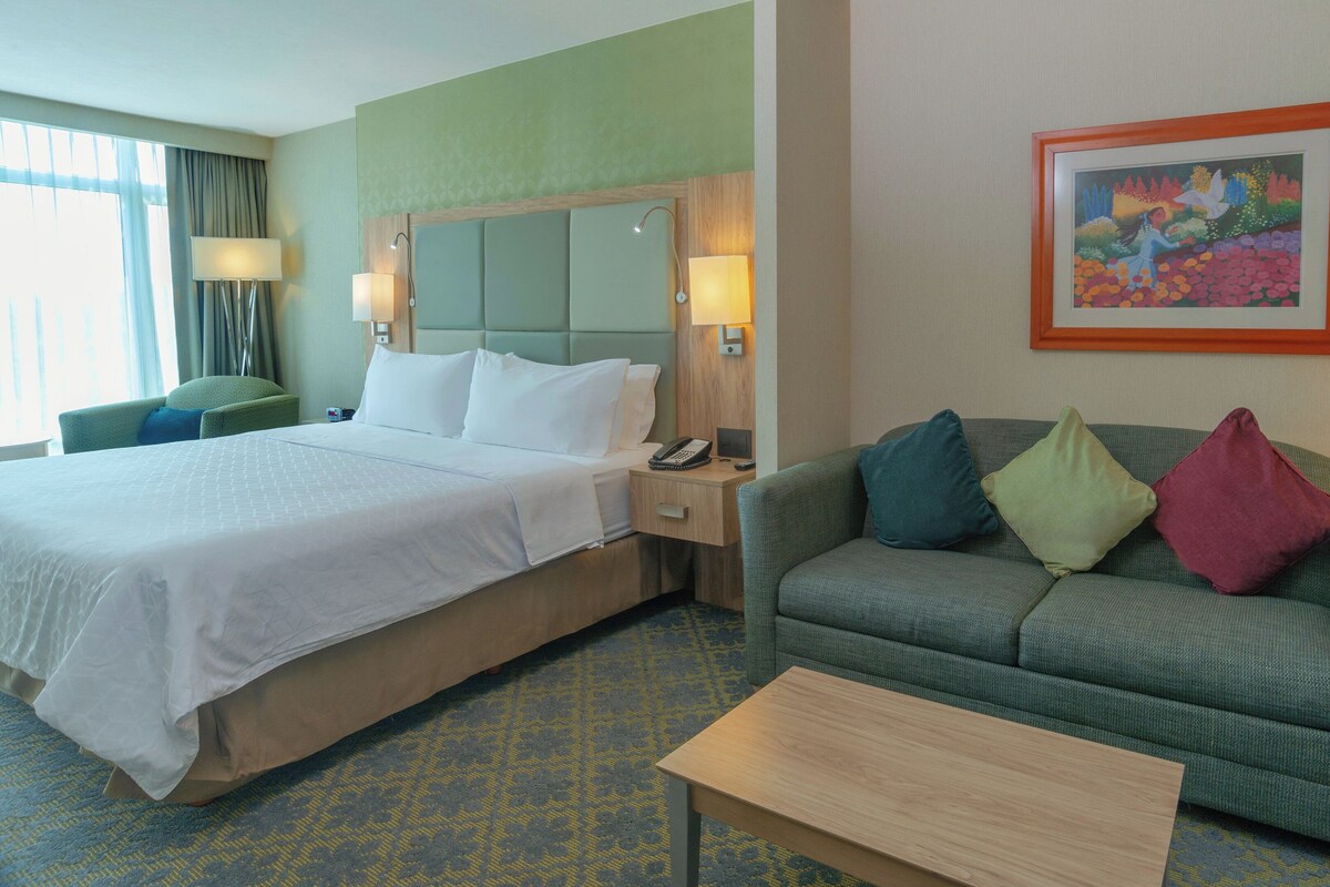A hotel room with a bed, a sofa, a coffee table, a painting, and a lamp.