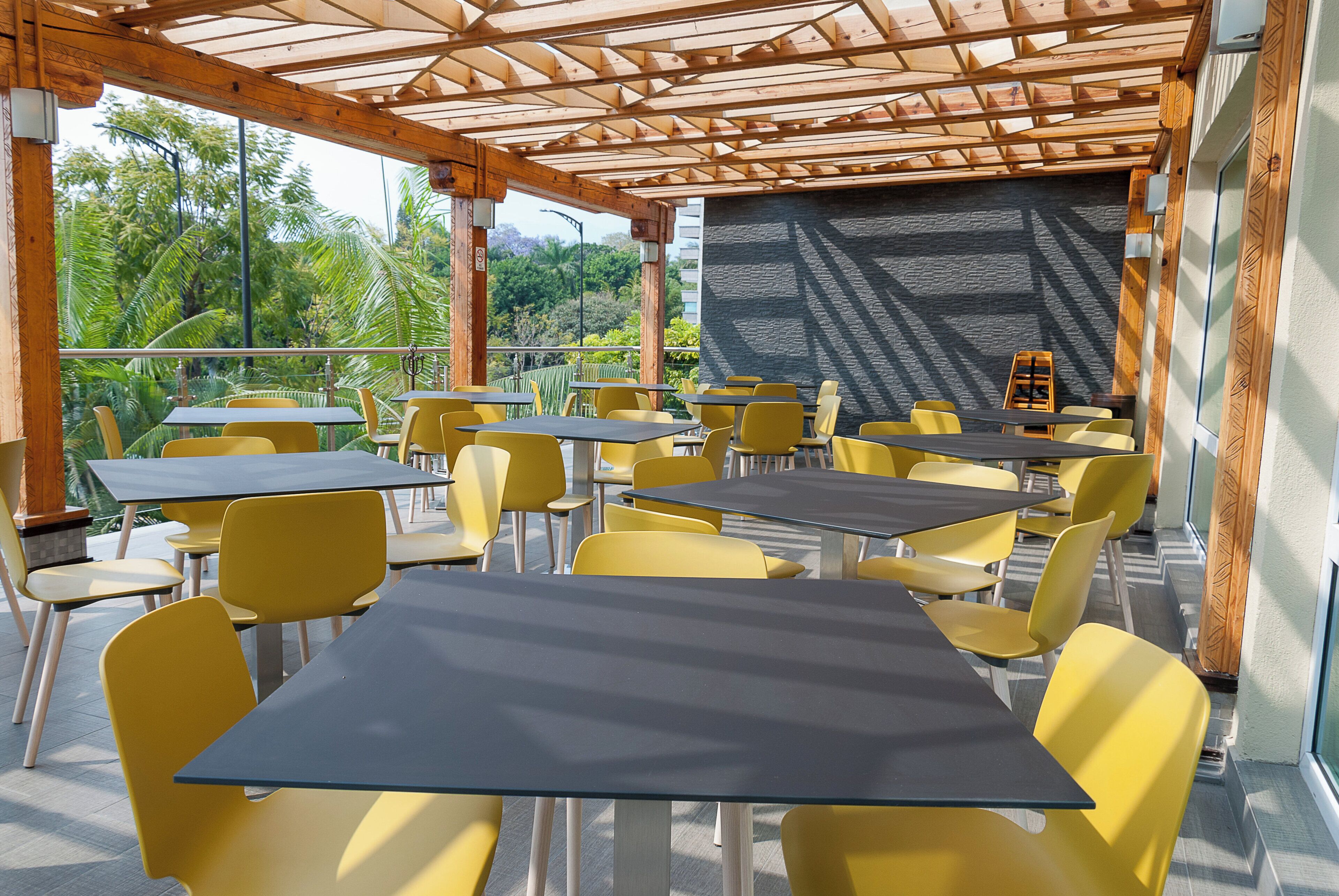 An outdoor cafe with tables and chairs, a wooden pergola, and a view of greenery.