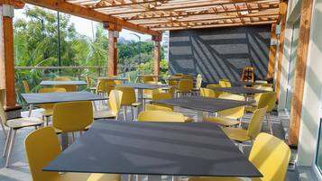 An outdoor cafe with tables and chairs, a wooden pergola, and a view of greenery.