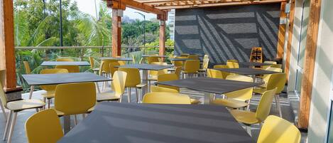 An outdoor cafe with tables and chairs, a wooden pergola, and a view of greenery.