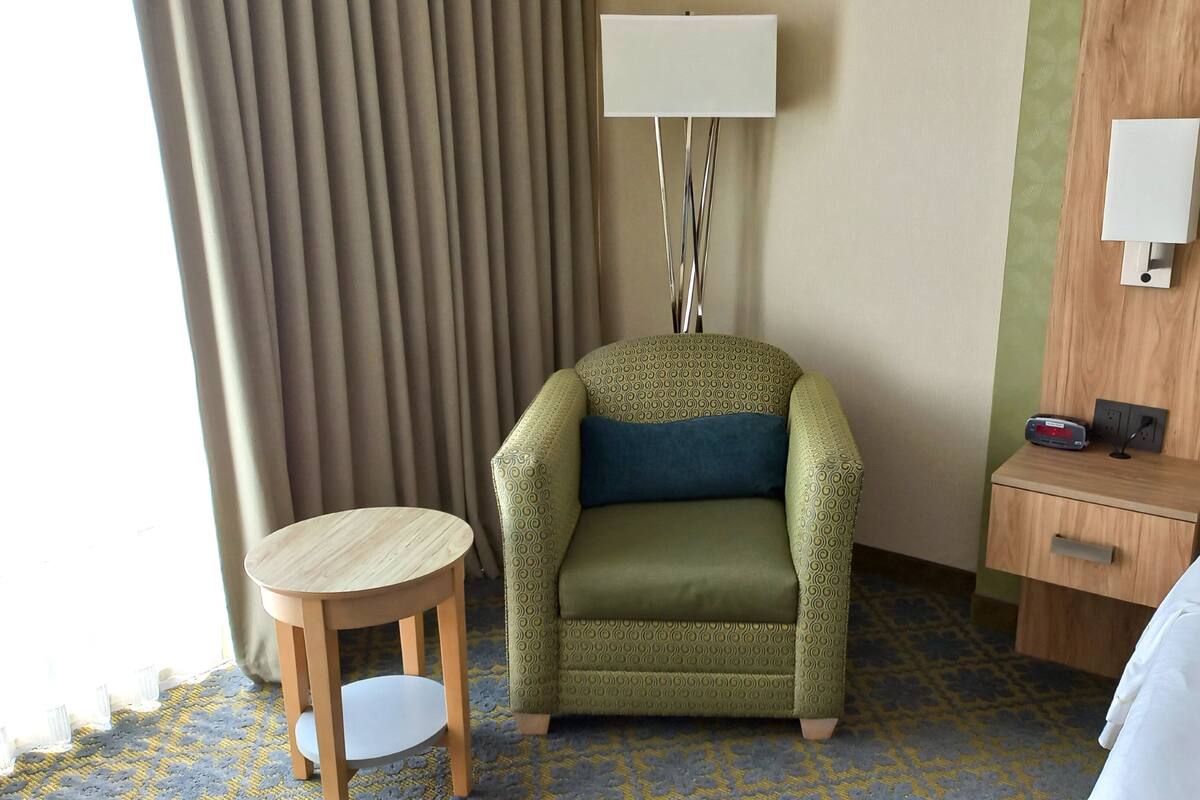 A hotel room with a green patterned armchair, a small wooden table, a bedside table with a lamp, and a bed with white linens.