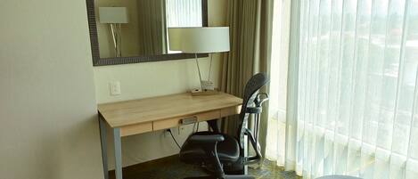 A hotel room with a wooden desk, a black office chair, a small table, a lamp, and a window with curtains.