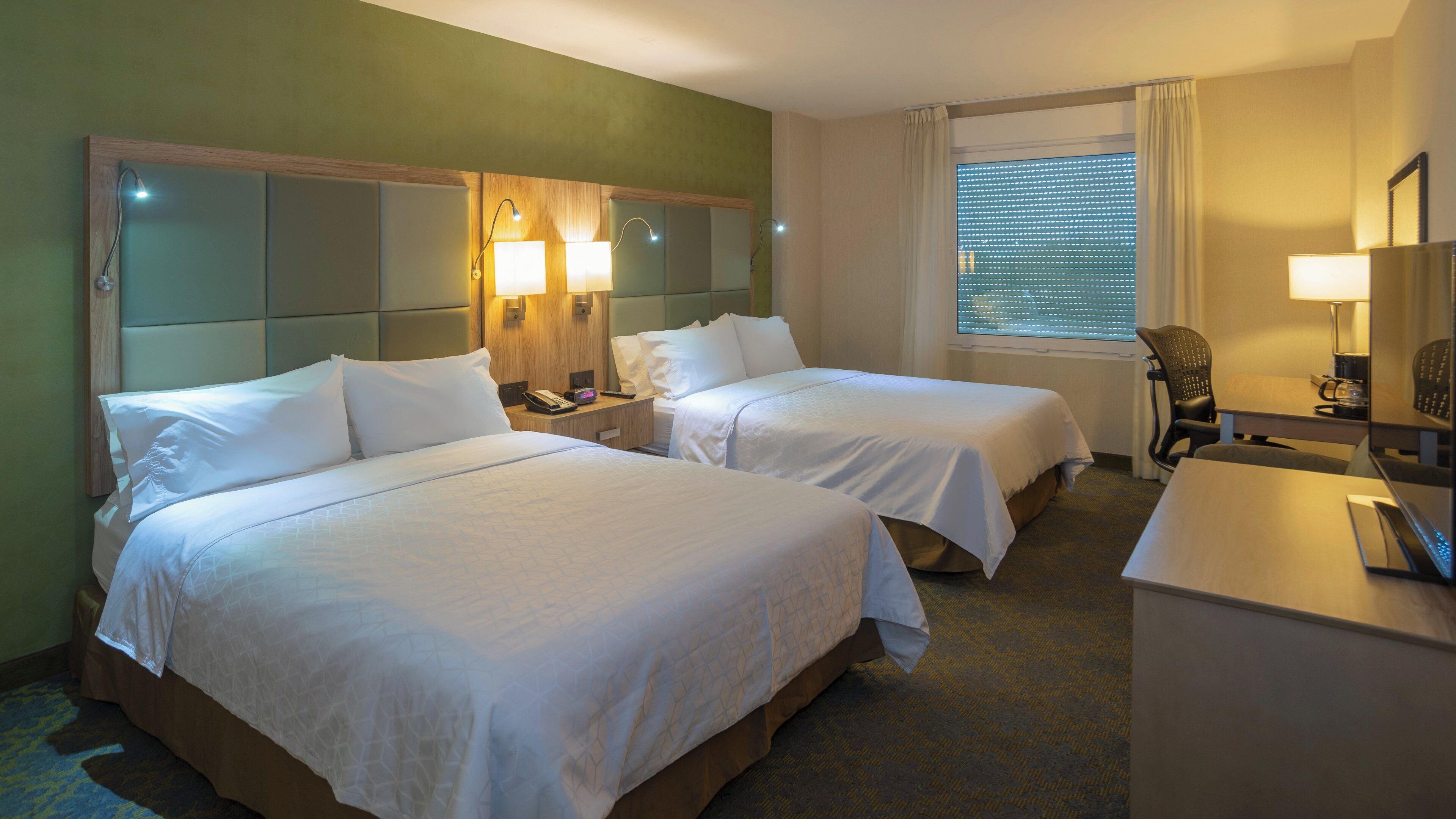 A hotel room with two beds, a desk, a chair, a lamp, and a window with blinds.
