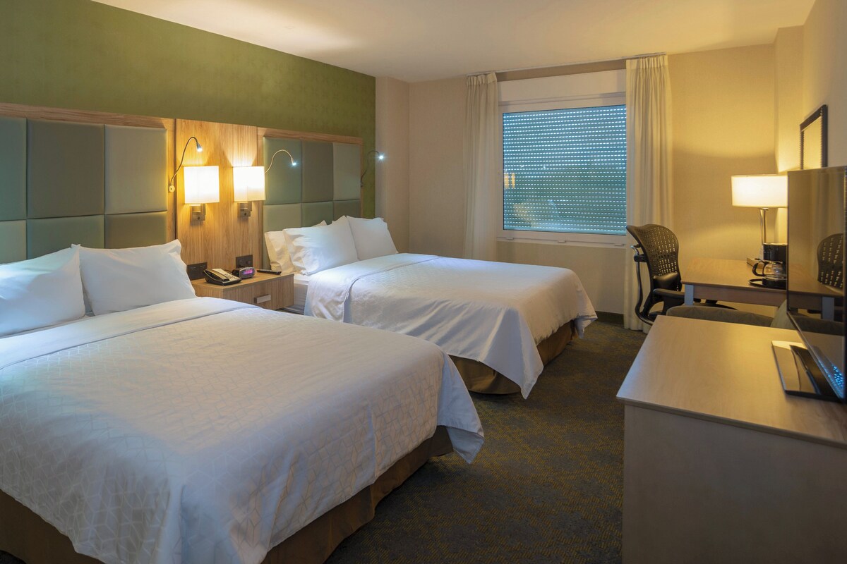 A hotel room with two beds, a desk, a chair, a lamp, and a window with blinds.