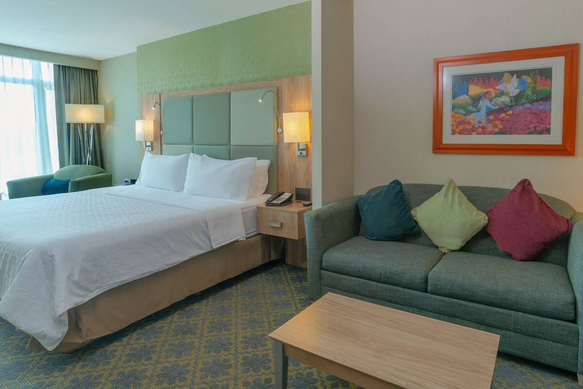 A hotel room with a bed, a sofa, a coffee table, a painting, and a lamp.