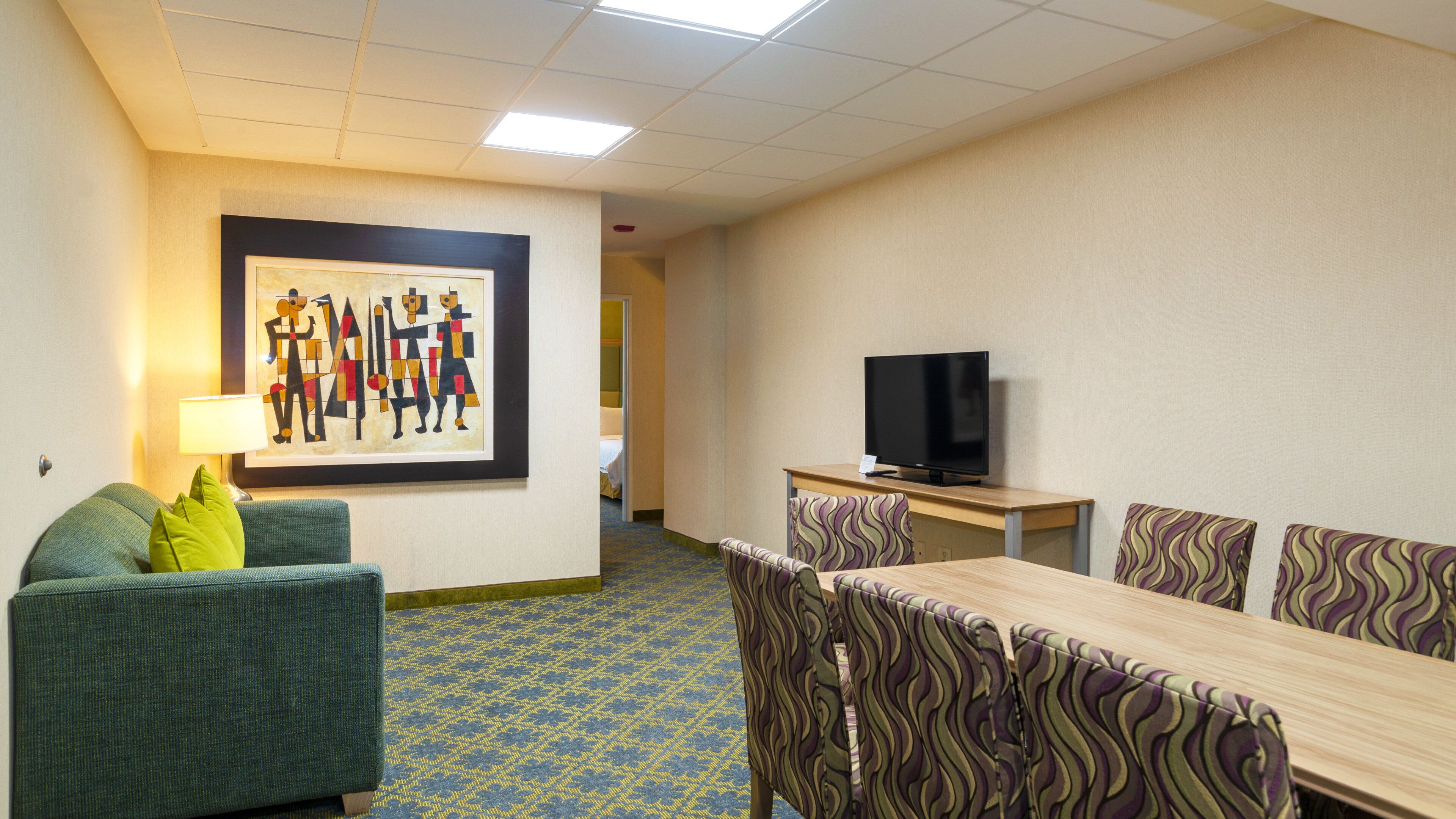 A hotel room with a sofa, a TV, a dining table with chairs, and a framed artwork on the wall.